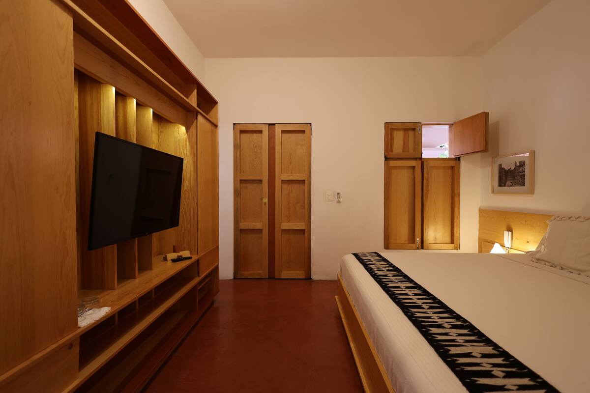 A bedroom with a TV mounted on a wooden cabinet, a bed with a patterned bedspread, and a nightstand with a lamp.