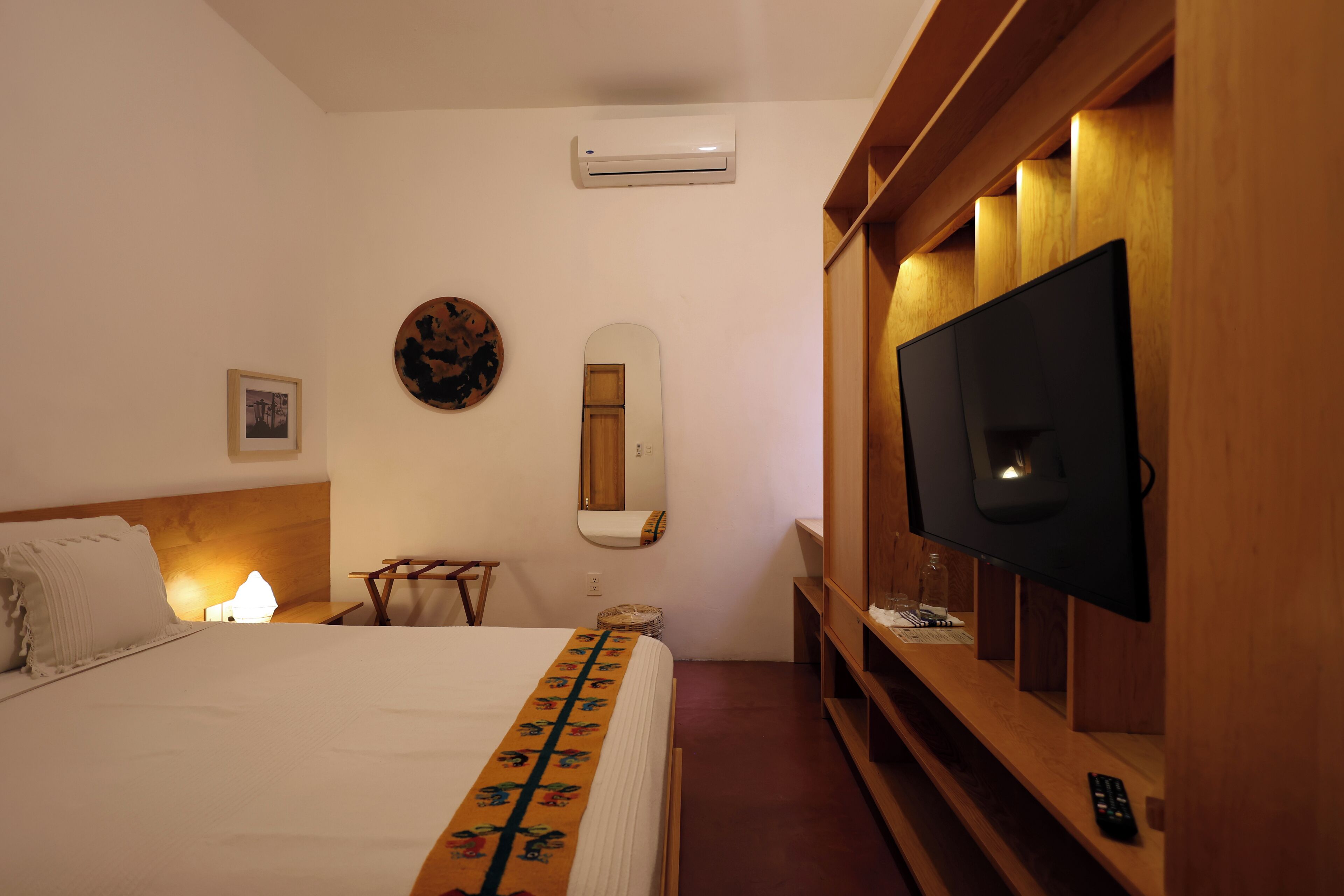 A hotel room with a bed, a television, a mirror, and a wall-mounted air conditioner.