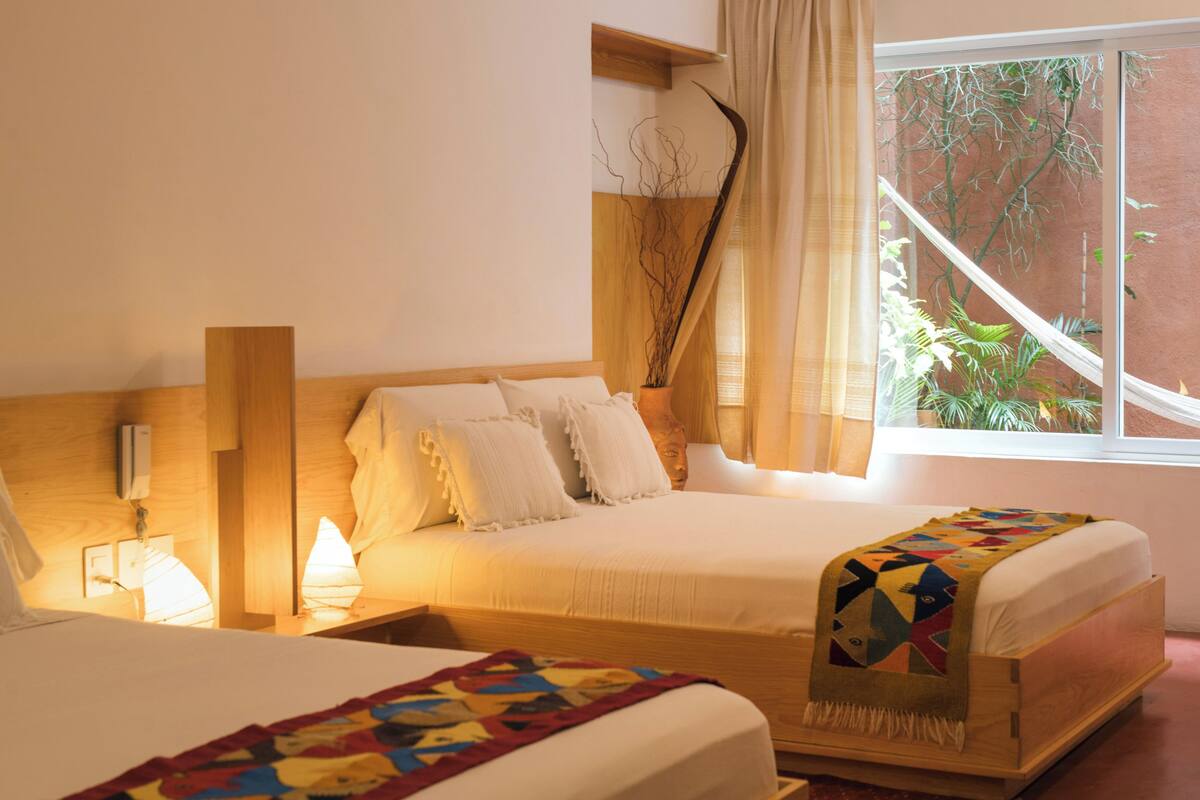 A bedroom with a wooden headboard, a bed with white bedding, a bedside table with a lamp, and a window with a view of plants.
