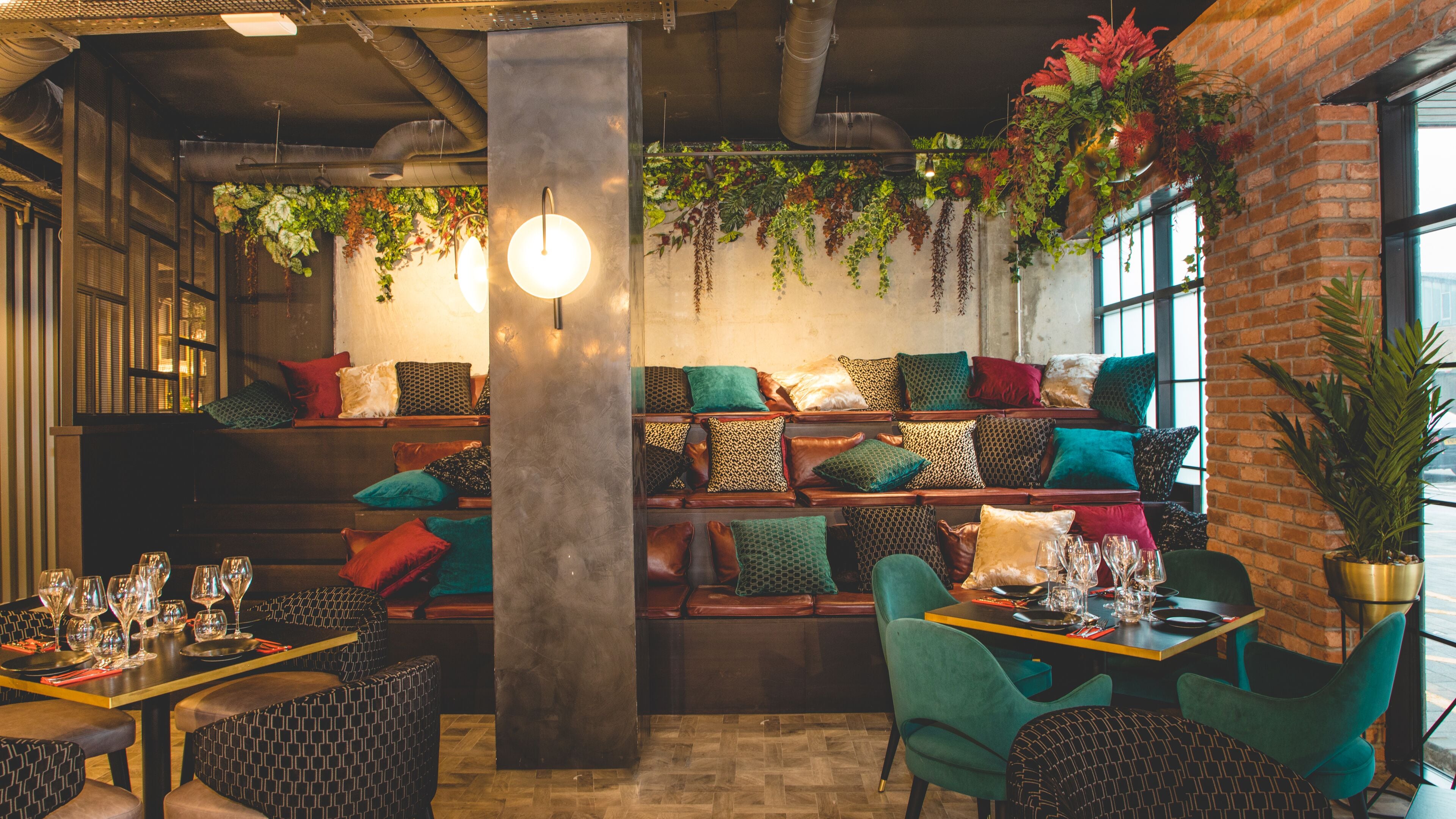A cozy cafe interior with wooden tables, cushioned seating, and hanging plants.