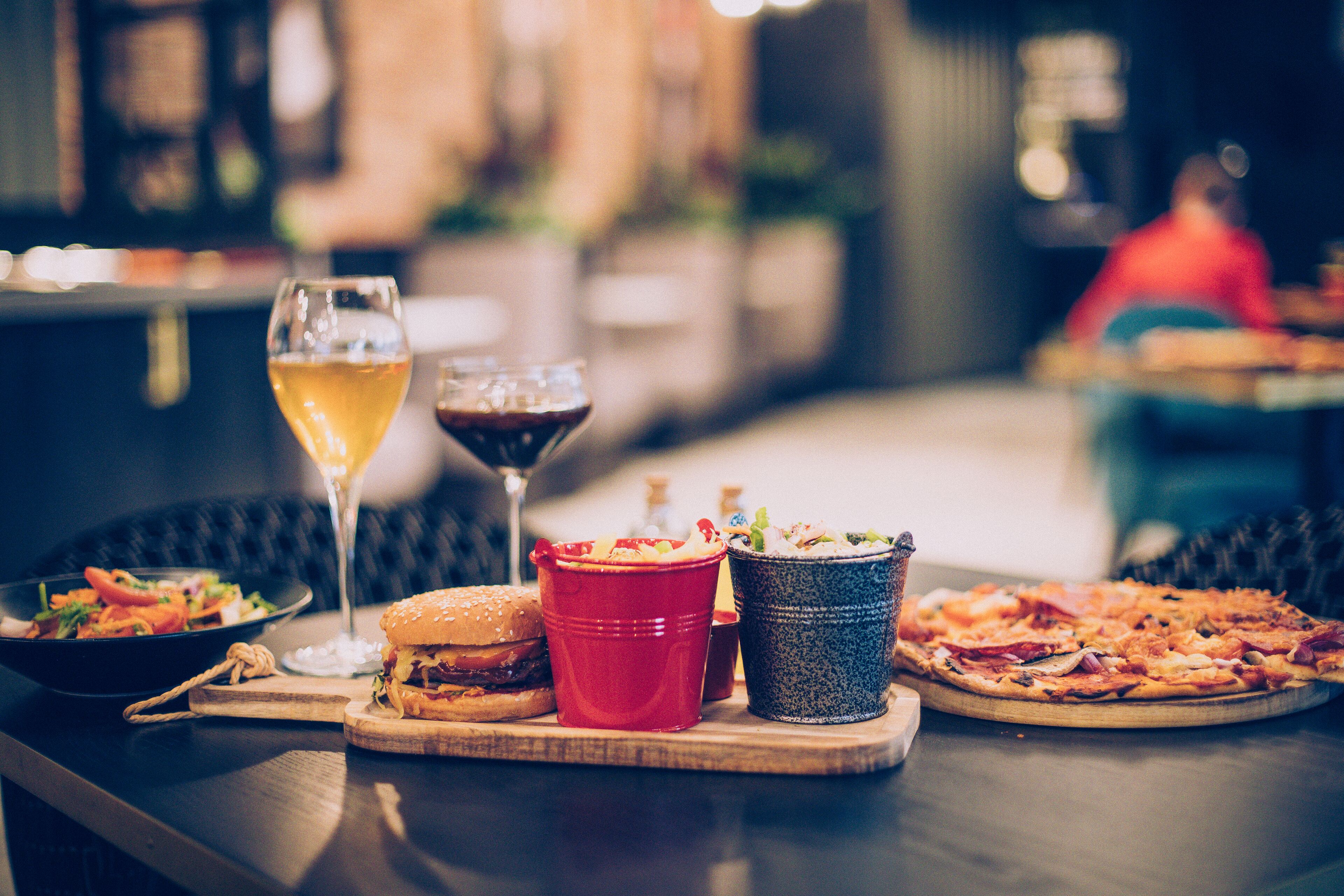 A dining table with a burger, a glass of wine, a pizza, and a bowl of salad.