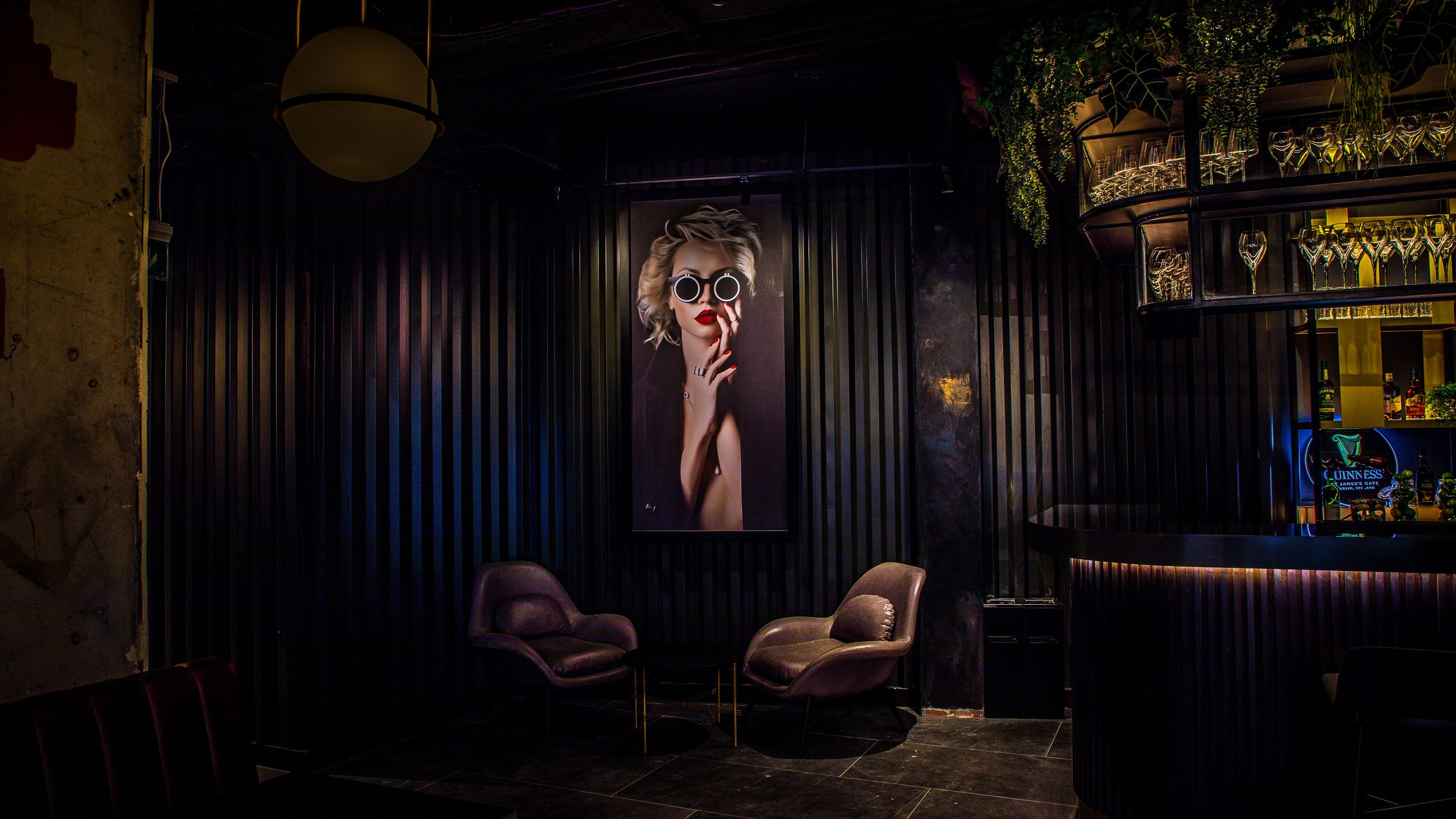 A dimly lit bar area with two leather chairs, a large framed portrait, and a bar stocked with glasses and bottles.