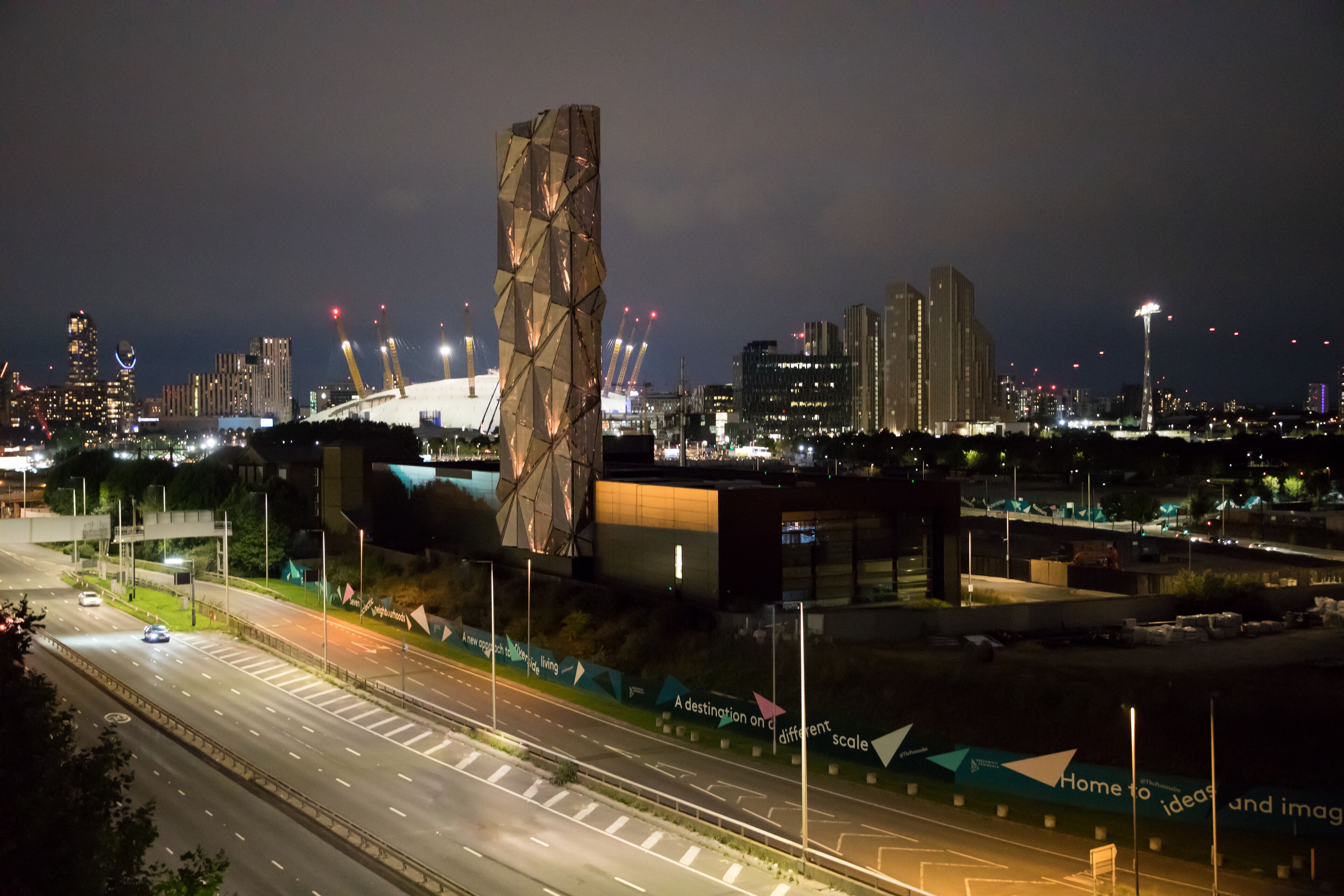 A nighttime cityscape with modern buildings, a prominent tower, and a well-lit road with a green area and a banner.