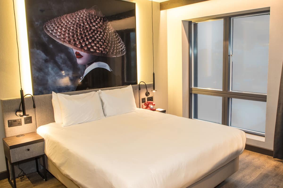Habitación de hotel con una cama grande, una mesita de noche con lámpara, un teléfono rojo y una ventana con cortinas.