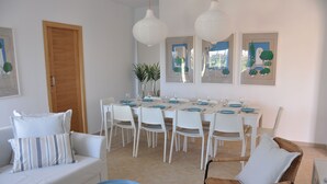 A dining area with a long table set for six, surrounded by white chairs, a wicker chair, and a small round table. - ECO Villa Nazaret with Solar Energy (Antigua)