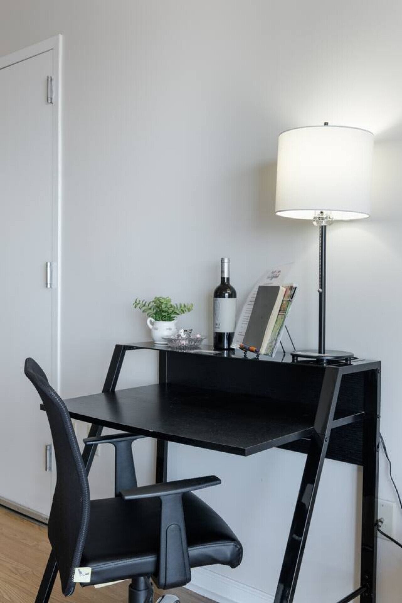 A compact workspace with a black desk, a chair, a floor lamp, a small potted plant, a wine bottle, and a magazine rack.
