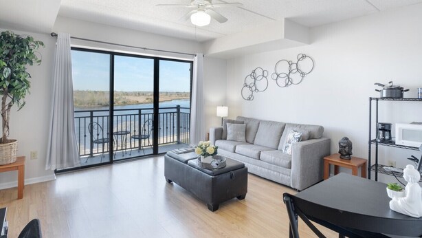A spacious living room with a grey sofa, a black ottoman, and a dining table. A large window offers a view of a body of water.
