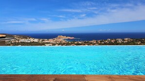 Una terraza de madera que da a una piscina azul cristalina, con vistas a un pueblo costero y al mar.