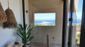 Una habitación con un ventanal amplio que ofrece vistas al mar y a un paisaje urbano. Hay un banco de madera, una planta en maceta y una lámpara colgante.