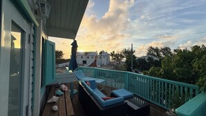 A wooden deck with a blue sofa and cushions, a table with seashells, and a view of buildings in the distance. - Beachside Studio with Car (Grand Turk)