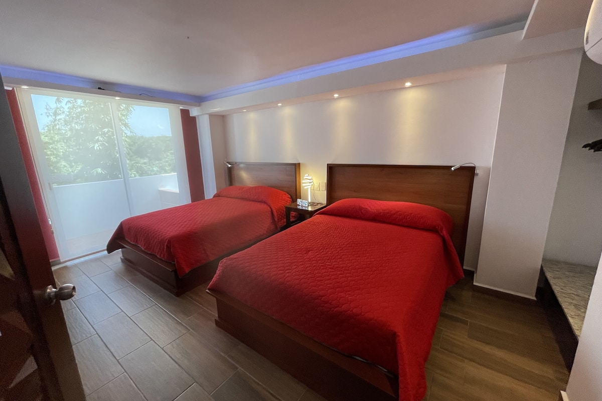 A hotel room with two beds, a wooden headboard, a nightstand with a lamp, and a view of greenery through a sliding door.