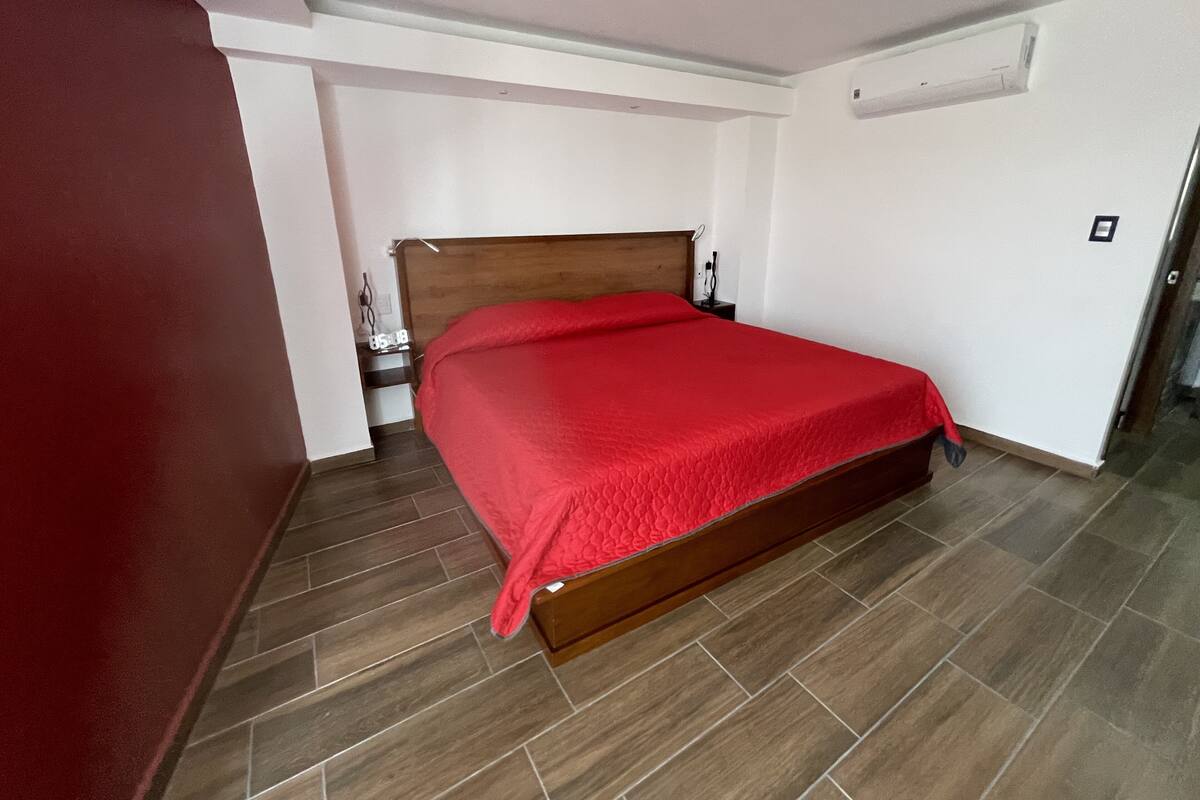 A bedroom with a wooden bed, a red bedspread, a nightstand, and a wall-mounted air conditioner.