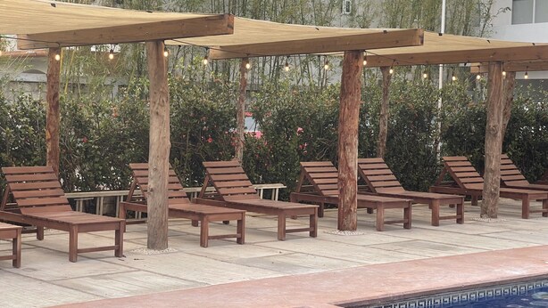 A poolside area with wooden lounge chairs and tables under a pergola, surrounded by greenery. - La Mansión del Faro (Tecolutla)