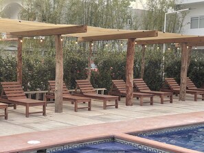 A poolside area with wooden lounge chairs and tables under a pergola, surrounded by greenery.