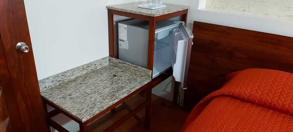 A compact room with a wooden bed, a small wooden table with a marble top, and a built-in refrigerator.