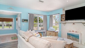 A room with a beige sofa, a small table, and a fireplace. There is a window with curtains and a painting above the fireplace. - Naut 2 Shabby Cottage-Awesome Pool Area, Pine Ave location close to everything (Anna Maria)