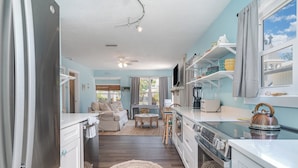 A modern kitchen with white cabinets, a stainless steel refrigerator, and a built-in oven. There is a dining area with a table and chairs, and a window with curtains. - Naut 2 Shabby Cottage-Awesome Pool Area, Pine Ave location close to everything (Anna Maria)