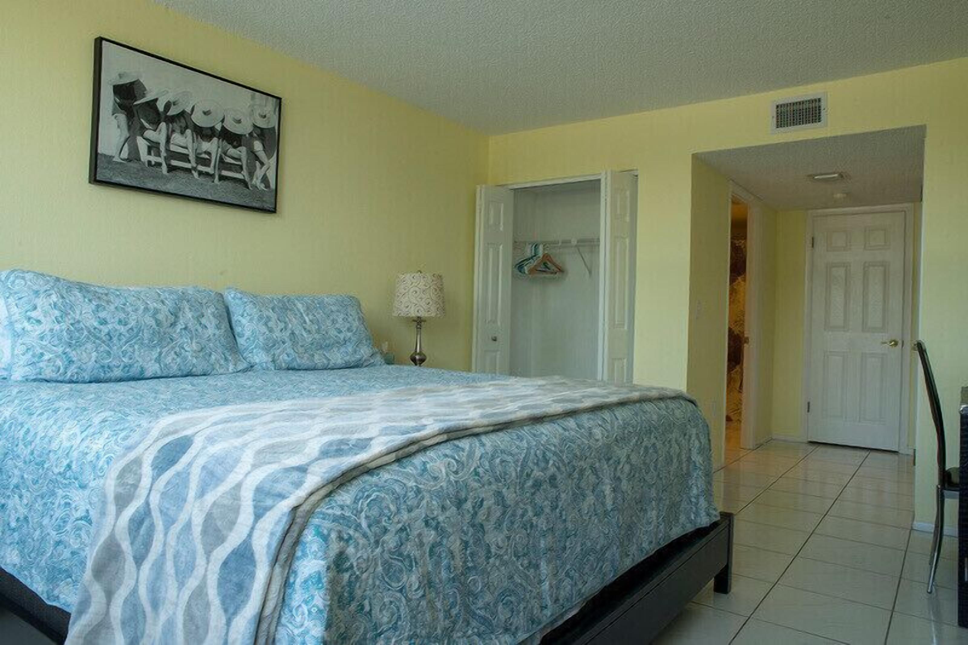 A bedroom with a bed, a nightstand, a lamp, a wardrobe, and a door leading to another room.
