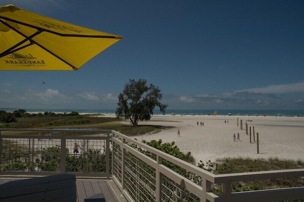 A beach view from a deck with a yellow umbrella, a tree, and people on the sand.