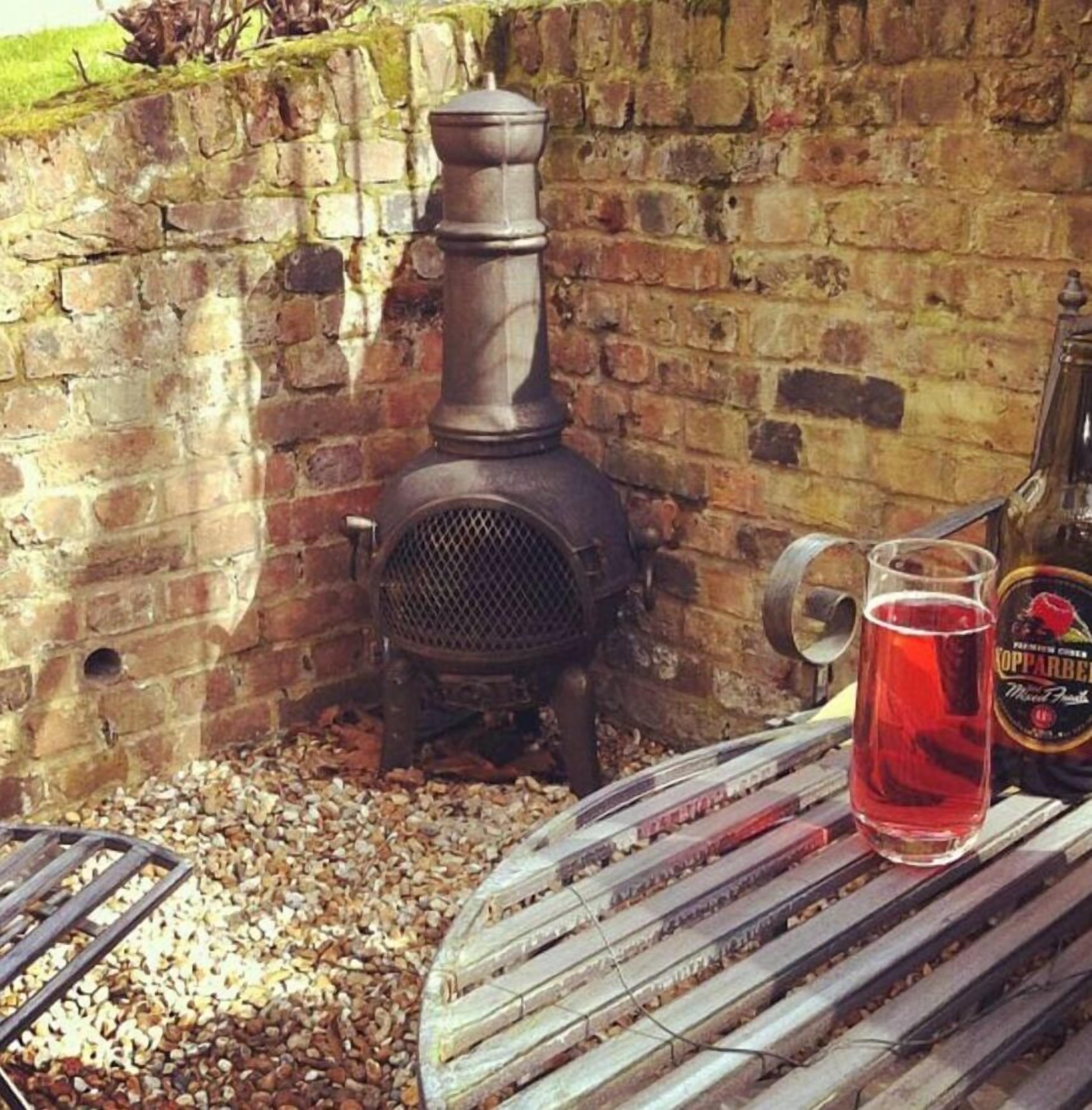 A rustic outdoor setting with a brick wall, a metal stove, and a glass of red beverage on a metal table.