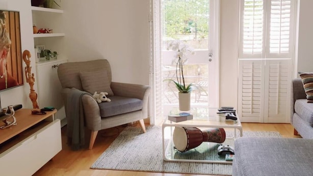 A living room with a grey sofa, a grey armchair, a coffee table with a drum, and a bookshelf with decorative items. - 2-Bed, Sleeps 4, Garden (London)