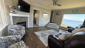 A living room with a fireplace, a TV, a ceiling fan, and a view of the ocean. - Spectacular views, close to down town Rockland (Owls Head)