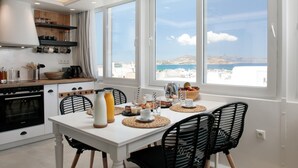 Une cuisine moderne avec des meubles blancs, une table à manger entourée de chaises noires et de grandes fenêtres offrant une vue sur la mer.