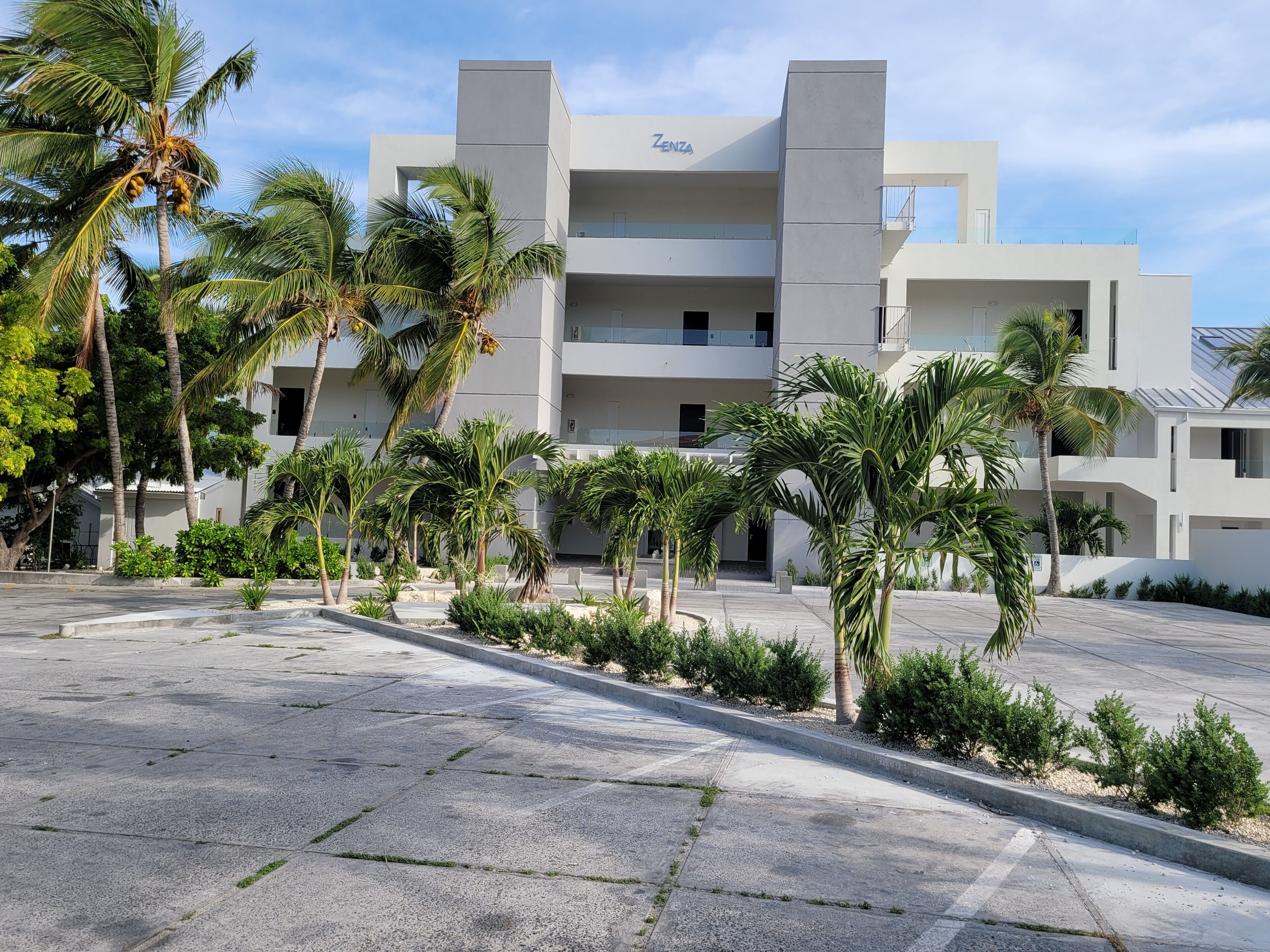 A modern building with the name 'Zenza' on the facade, surrounded by palm trees and a paved courtyard.