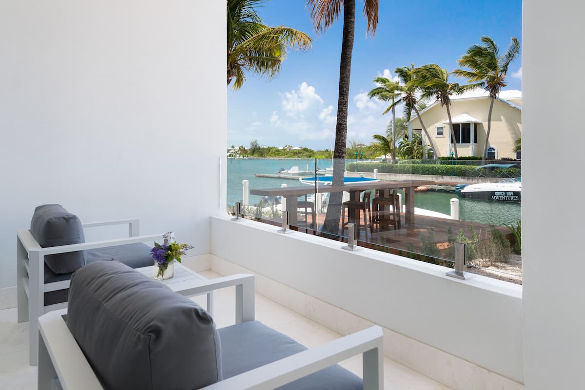A balcony with a white chair and a small table with a vase of flowers. A marina with boats and palm trees is visible in the background.