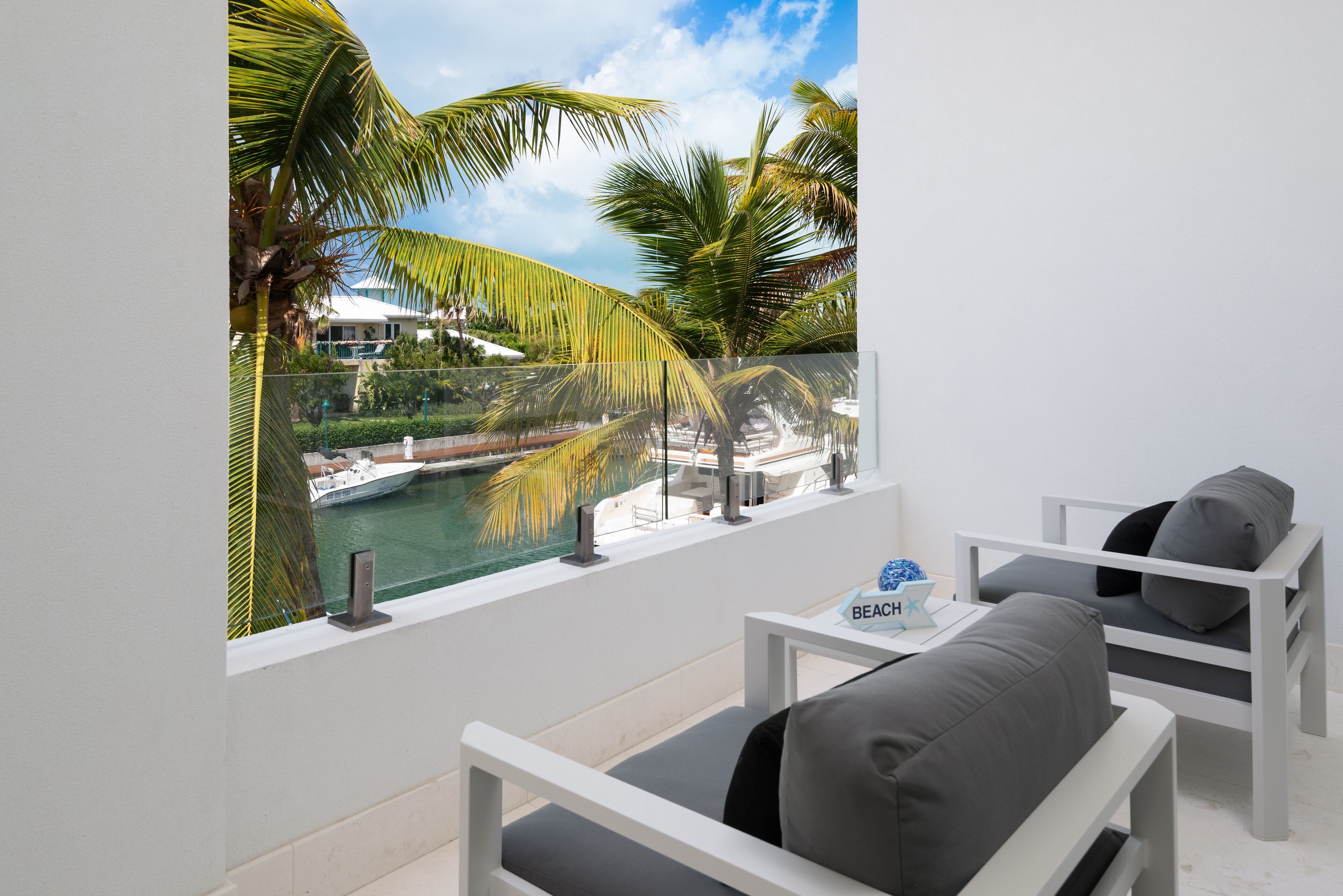A balcony with a view of a pool and palm trees, white furniture, and a sign that reads "BEACH".