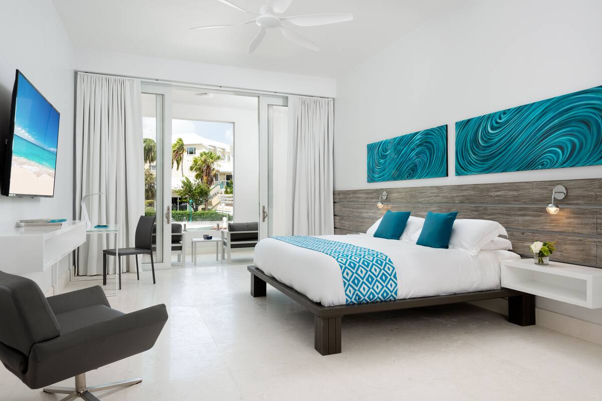 A modern hotel room with a bed, a desk, a chair, and a balcony with a view of palm trees.