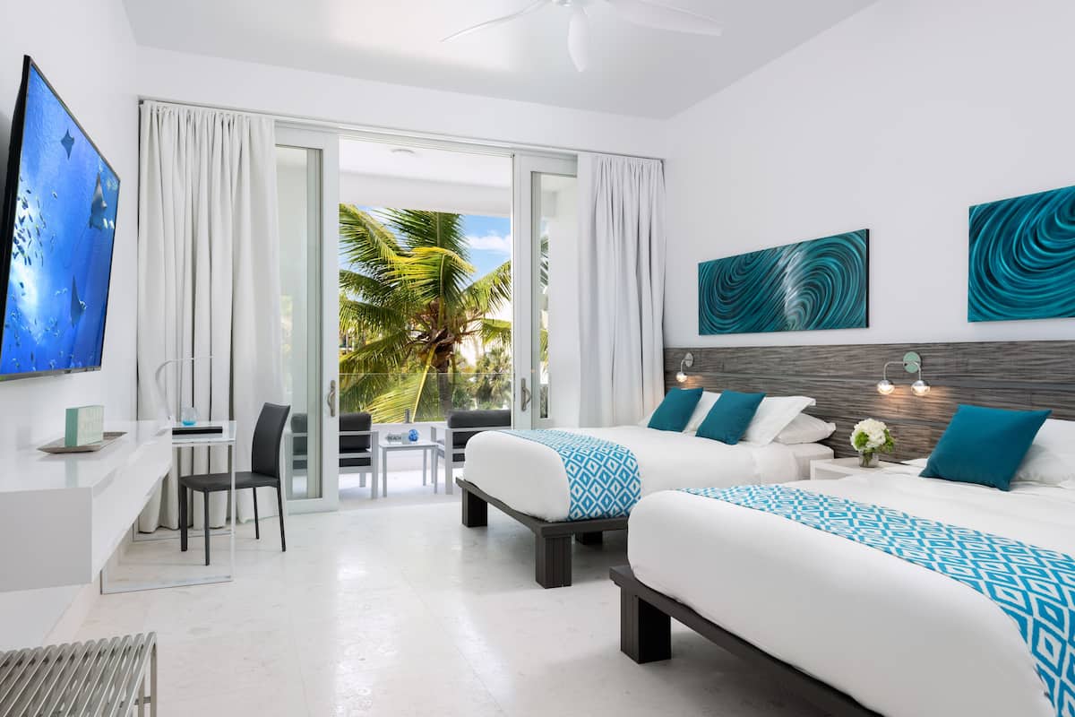 A modern hotel room with two beds, a desk, and a balcony with a view of palm trees.