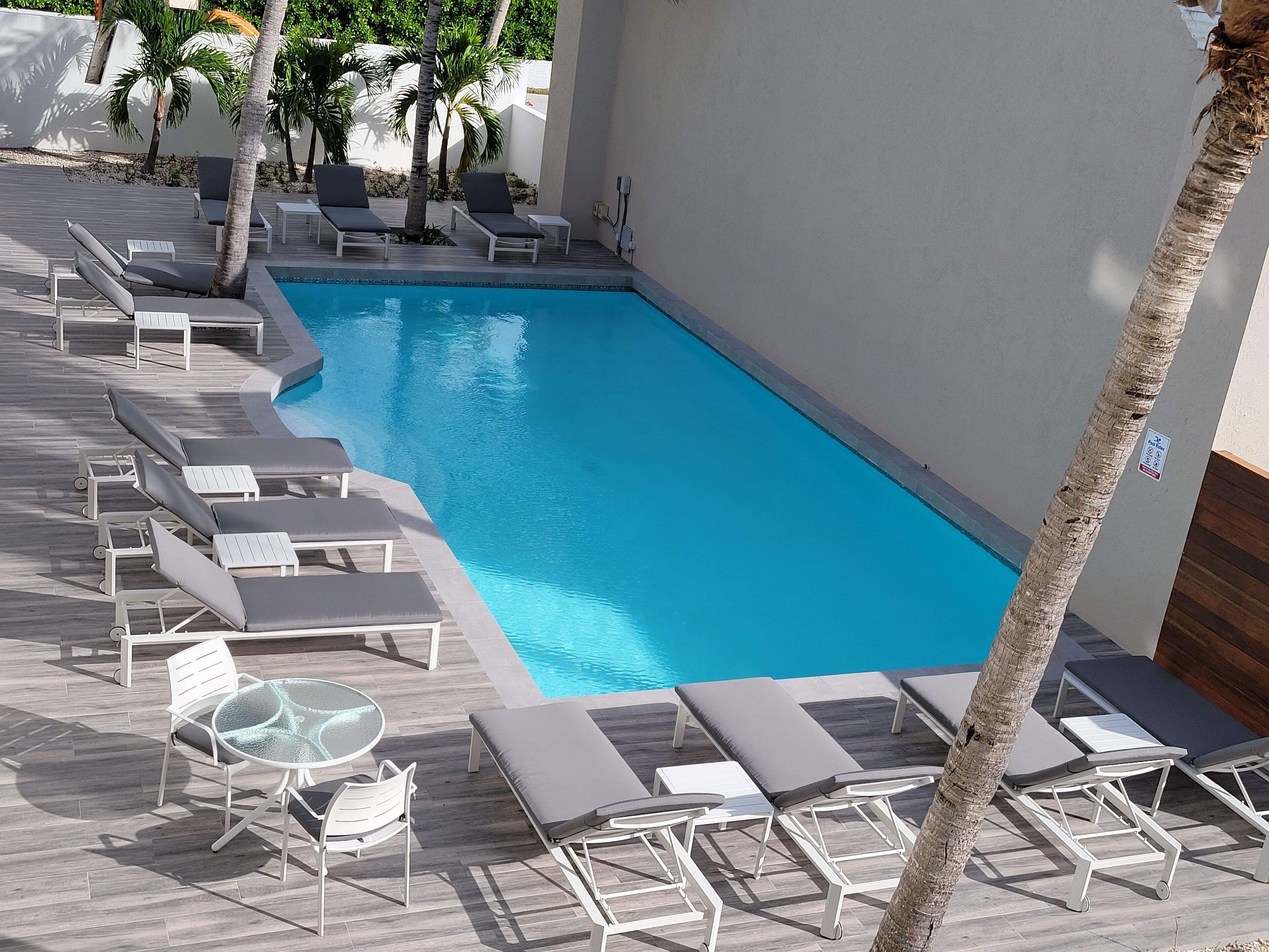 A rectangular swimming pool with clear blue water, surrounded by white lounge chairs and tables.