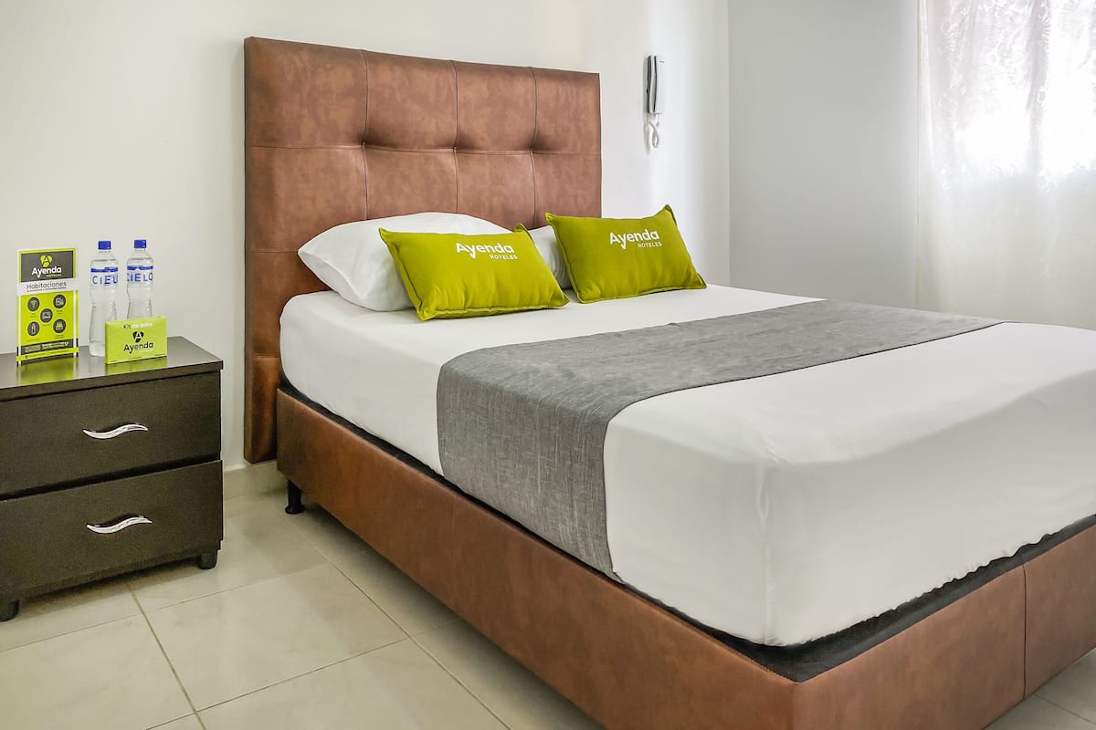 Ein Hotelzimmer mit braunem Kopfteil, weißer Bettwäsche und grünen Kissen mit dem Aufdruck "Ayenda Hotels". Auf einem Nachttisch stehen Toilettenartikel und auf dem Bett liegt eine graue Decke.