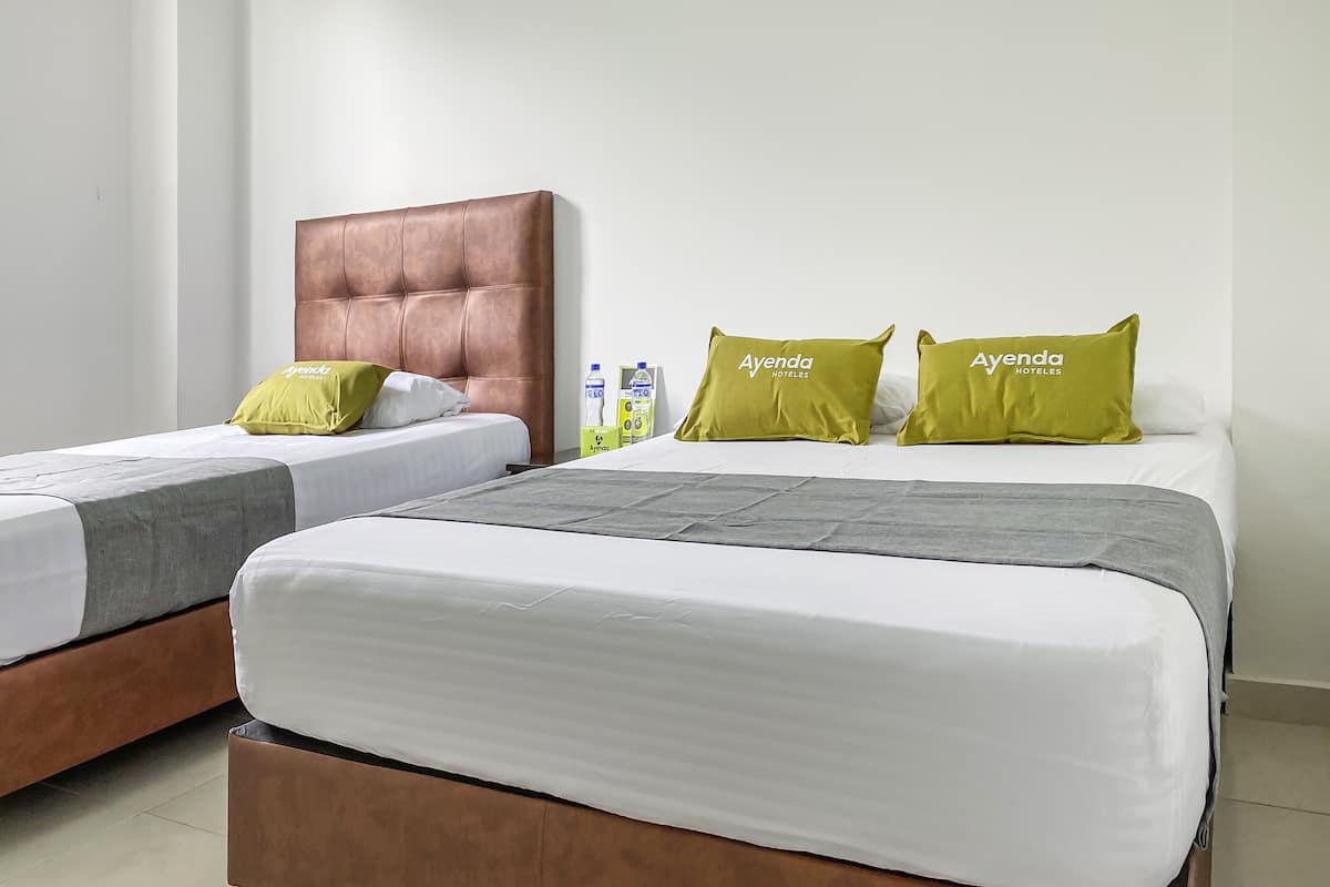 Ein Hotelzimmer mit zwei Betten, jeweils mit brauner Kopfteil und weißer Bettwäsche. Auf dem Nachttisch liegen grüne Kissen mit dem Logo des Hotels und eine Milchflasche.