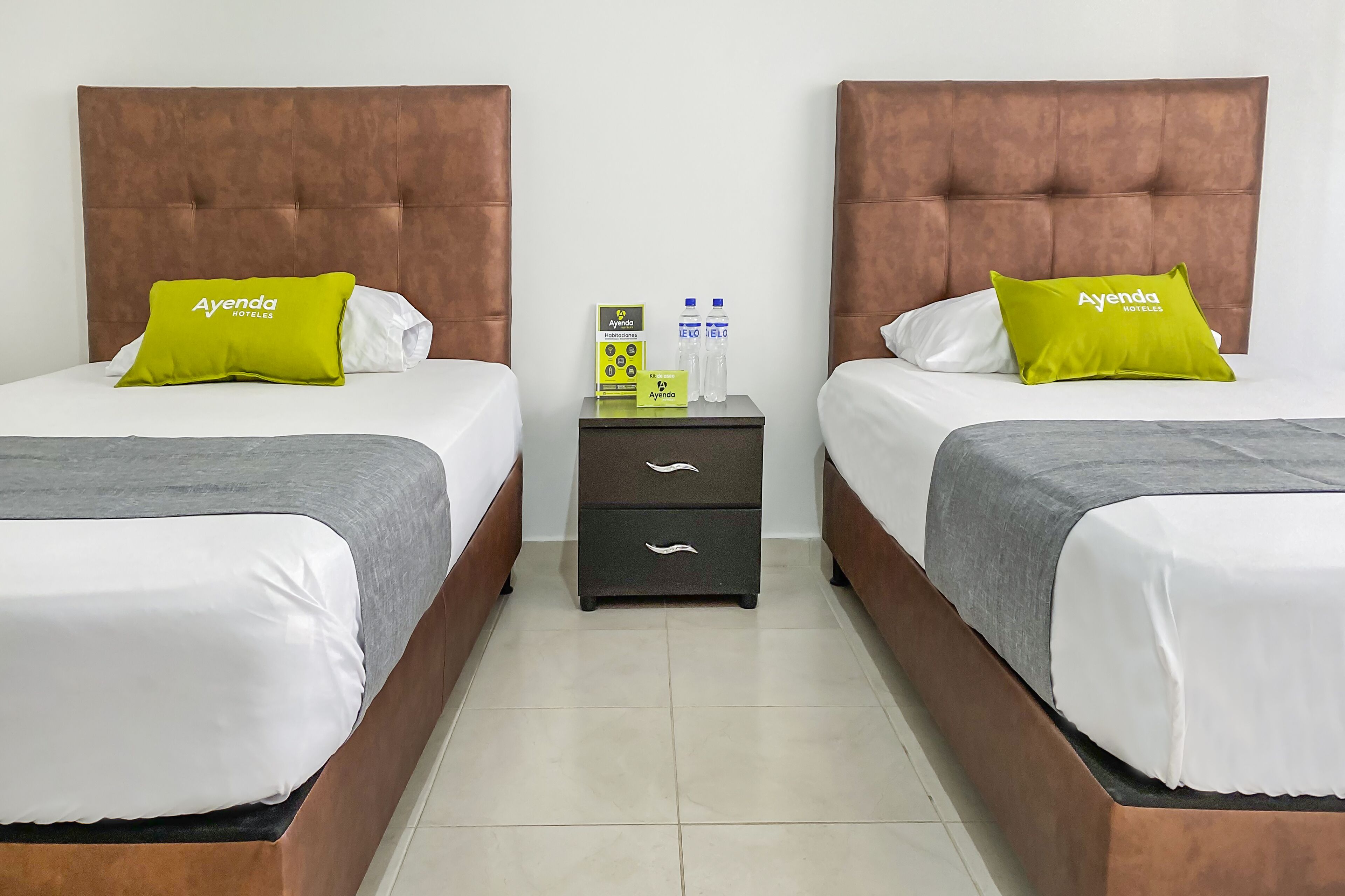 A hotel room with two beds, each with a brown headboard, white bedding, and a green pillow with "Ayenda Hotels" text. A nightstand with a drawer and two water bottles is between the beds.