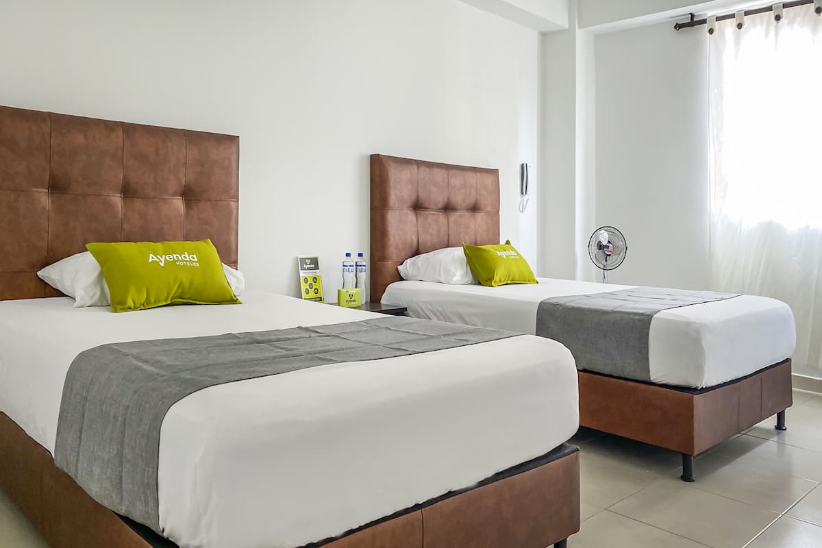 Zwei Einzelbetten mit braunen Kopfteilen, weißer Bettwäsche und grünen Kissen mit 'Ayenda Hotels'-Branding. Auf dem Nachttisch sind ein Ventilator und Toilettenartikel zu sehen.