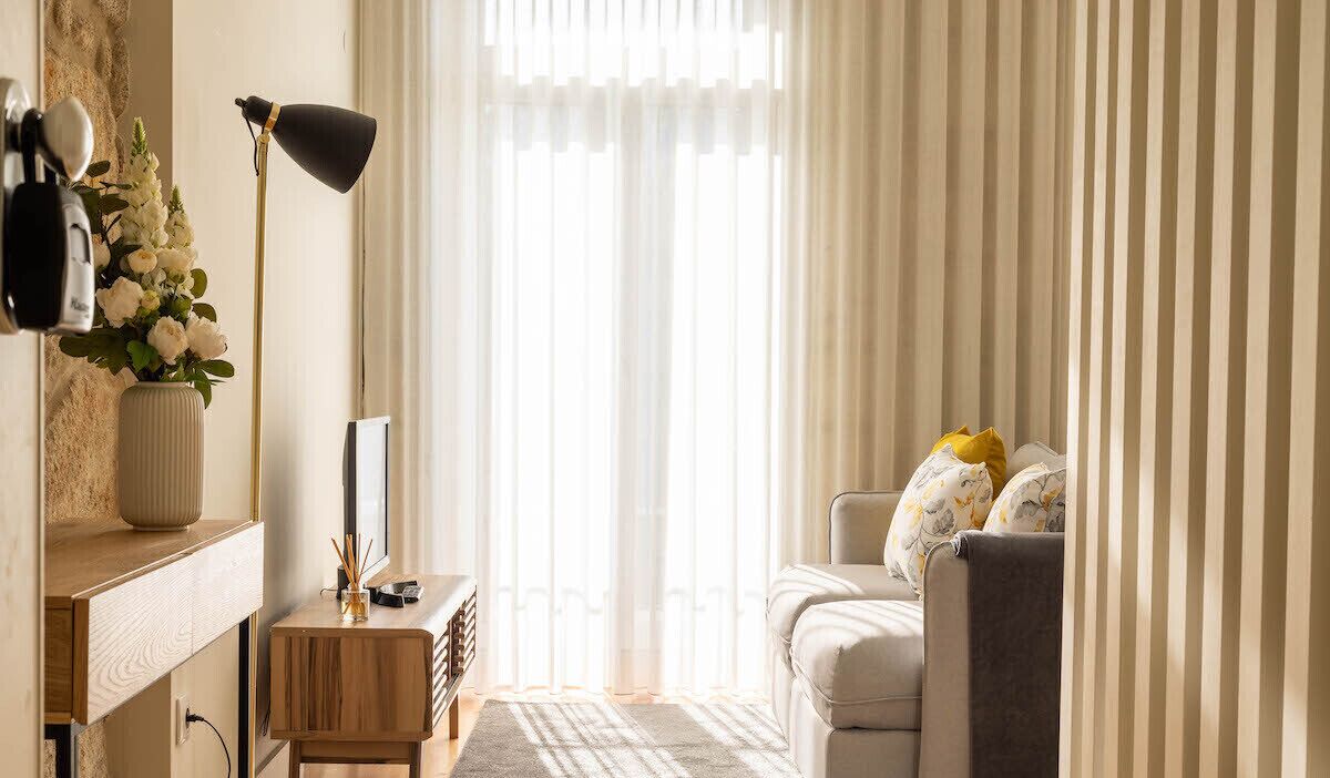 Ein modernes Wohnzimmer mit einer Couch, einer Lampe, einer Vase mit Blumen und einem Fenster mit durchsichtigen Vorhängen.