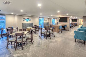 Un moderno vestíbulo de hotel con varias zonas de estar, incluyendo mesas, sillas, un bar y un escenario para presentaciones.