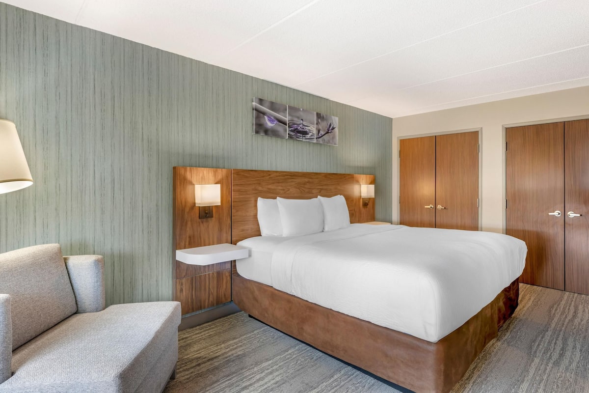 Hotellihuone, jossa on suuri sänky, puinen pääty, yöpöytä lampulla, tuoli ja seinälle ripustettu taideteos.
