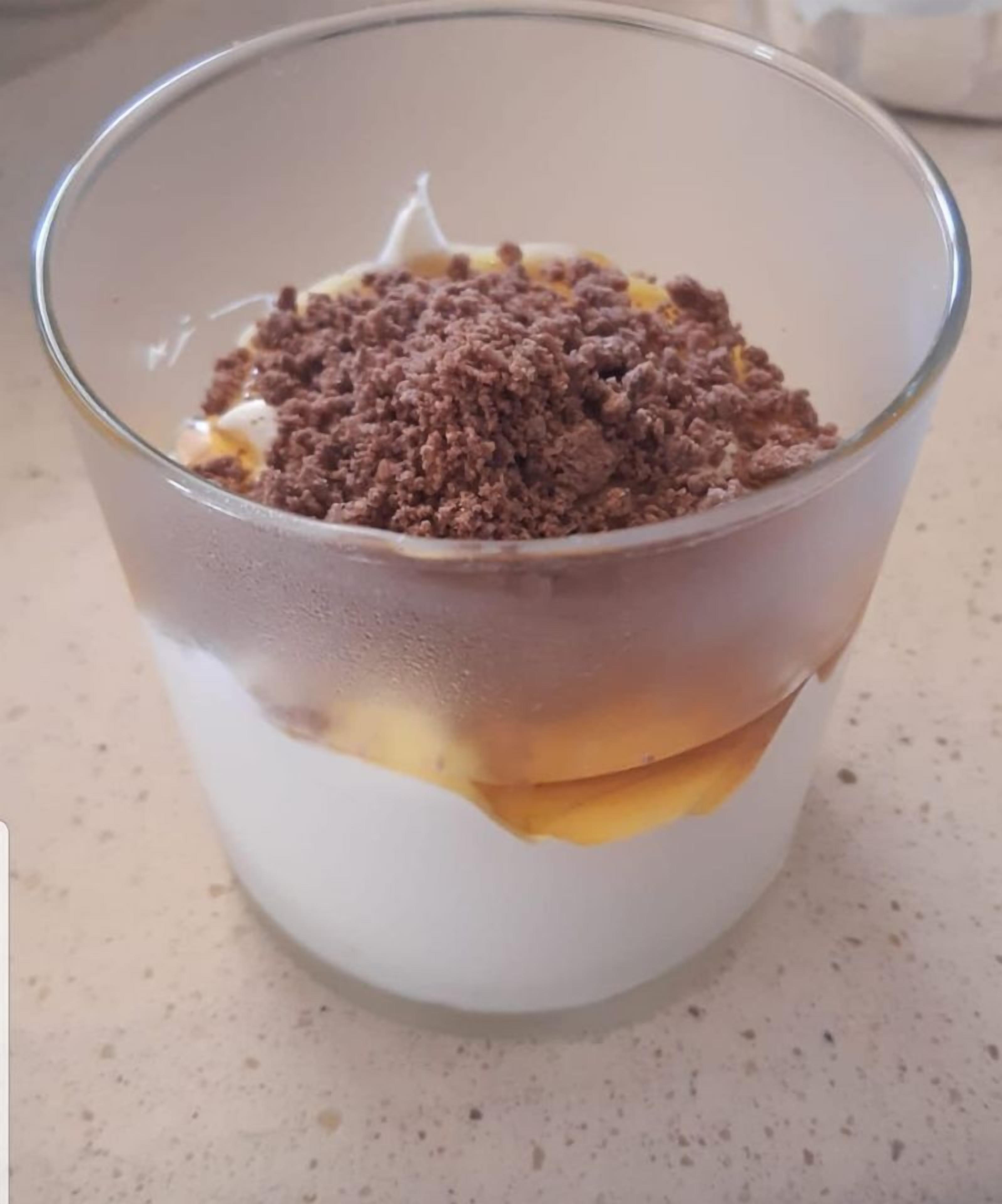 Un dessert servi dans un verre transparent, composé de plusieurs couches : une base blanche, une couche intermédiaire jaune et une garniture brune.