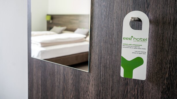 Habitación de hotel con puerta de madera y espejo que refleja una cama bien tendida con sábanas y almohadas blancas.