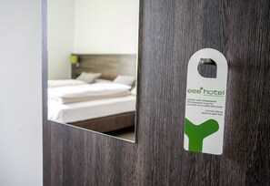 A hotel room with a wooden door and a mirror reflecting a neatly made bed with white linens and pillows. - eee hotel Graz (Seiersberg-Pirka)