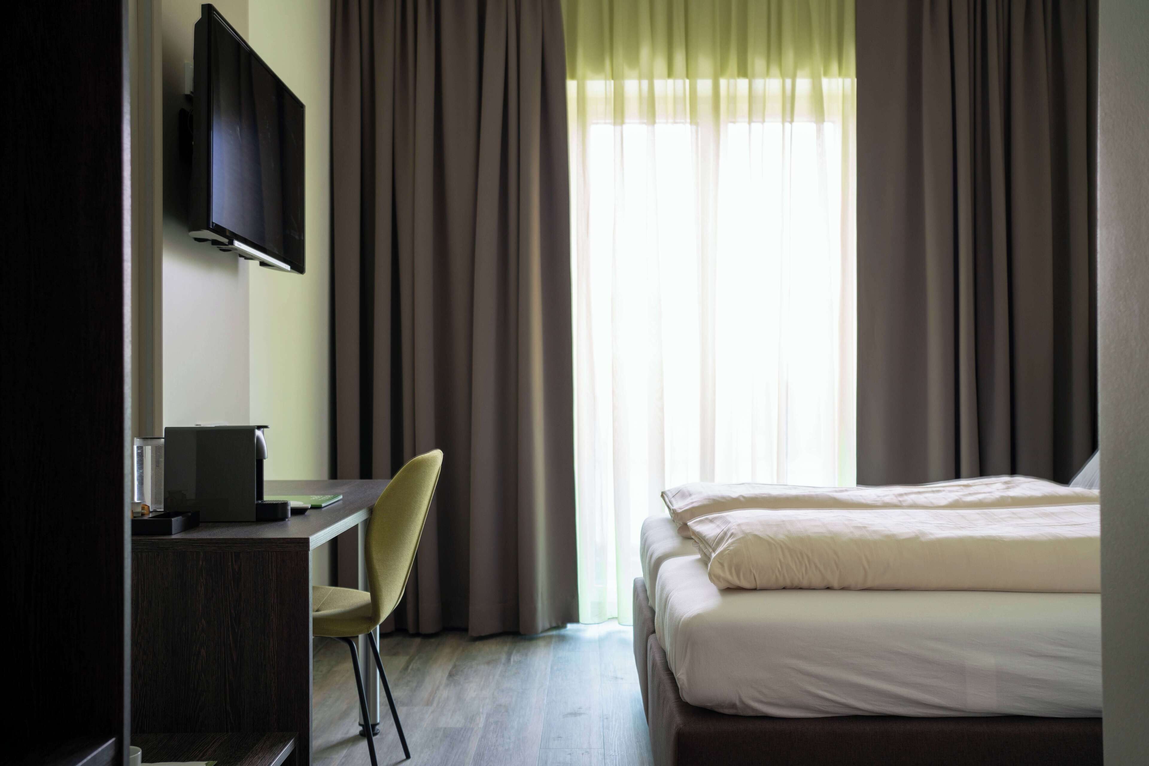 Ein Hotelzimmer mit Bett, Schreibtisch mit Stuhl, Fernseher und einem Fenster mit Vorhängen.