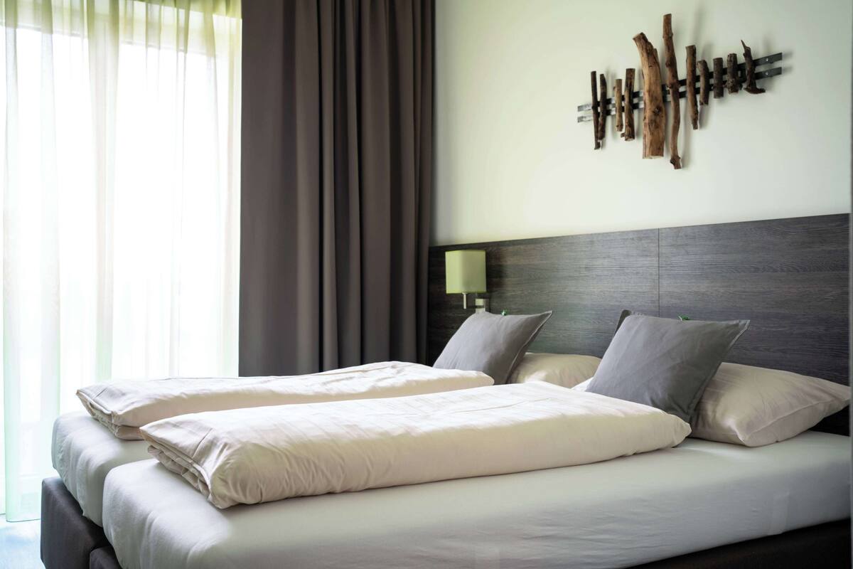Quarto de hotel com duas camas, cabeceira de madeira, luminária fixada na parede e vista para o exterior através de cortinas finas.