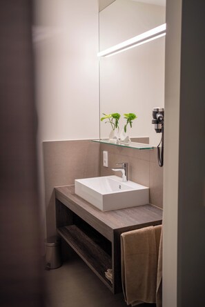 A modern bathroom with a white sink, a glass shelf with plants, a hairdryer, and a towel hanging on the wall.