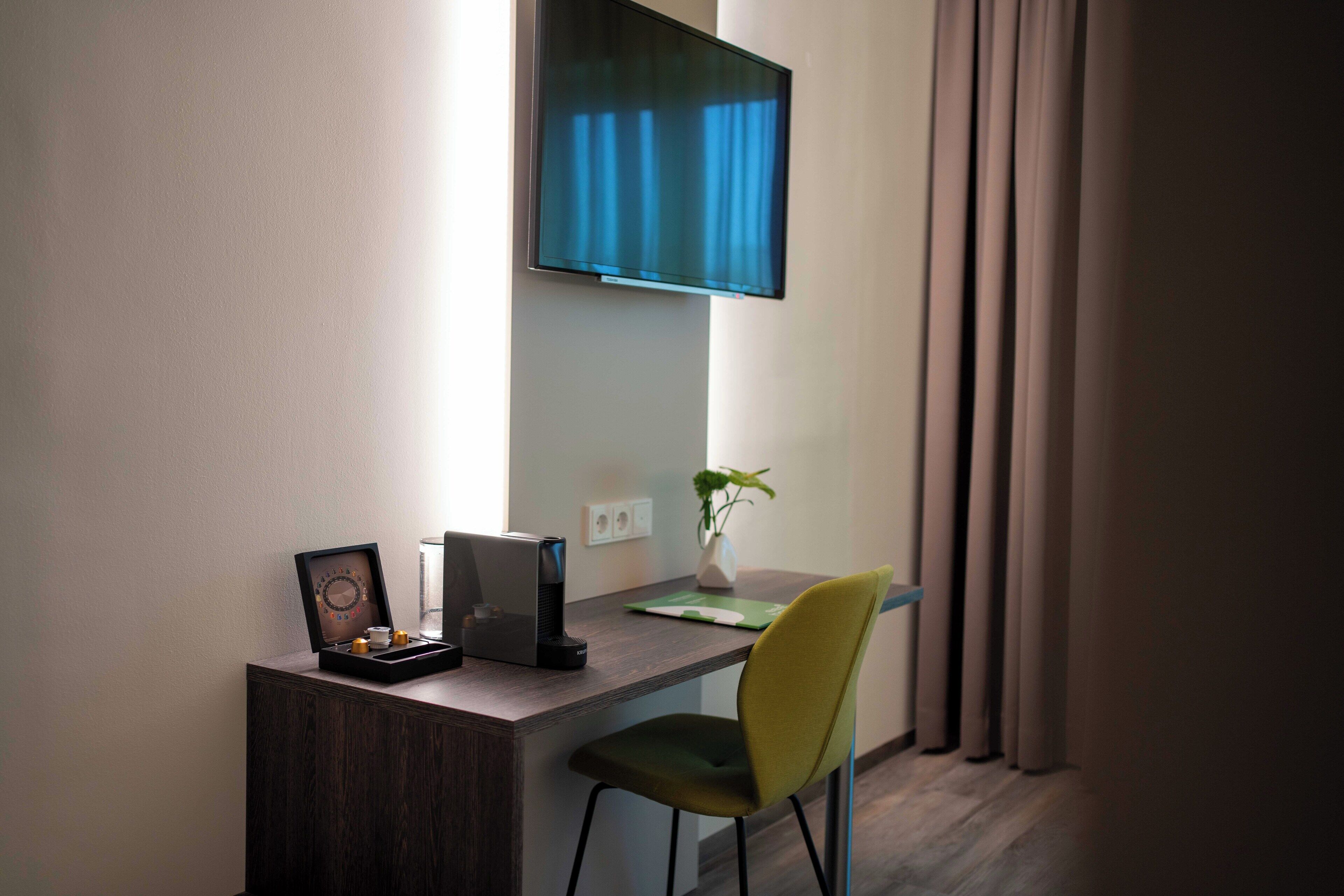 A modern hotel room with a desk, a flat-screen TV mounted on the wall, a green chair, a small plant, and a framed picture.