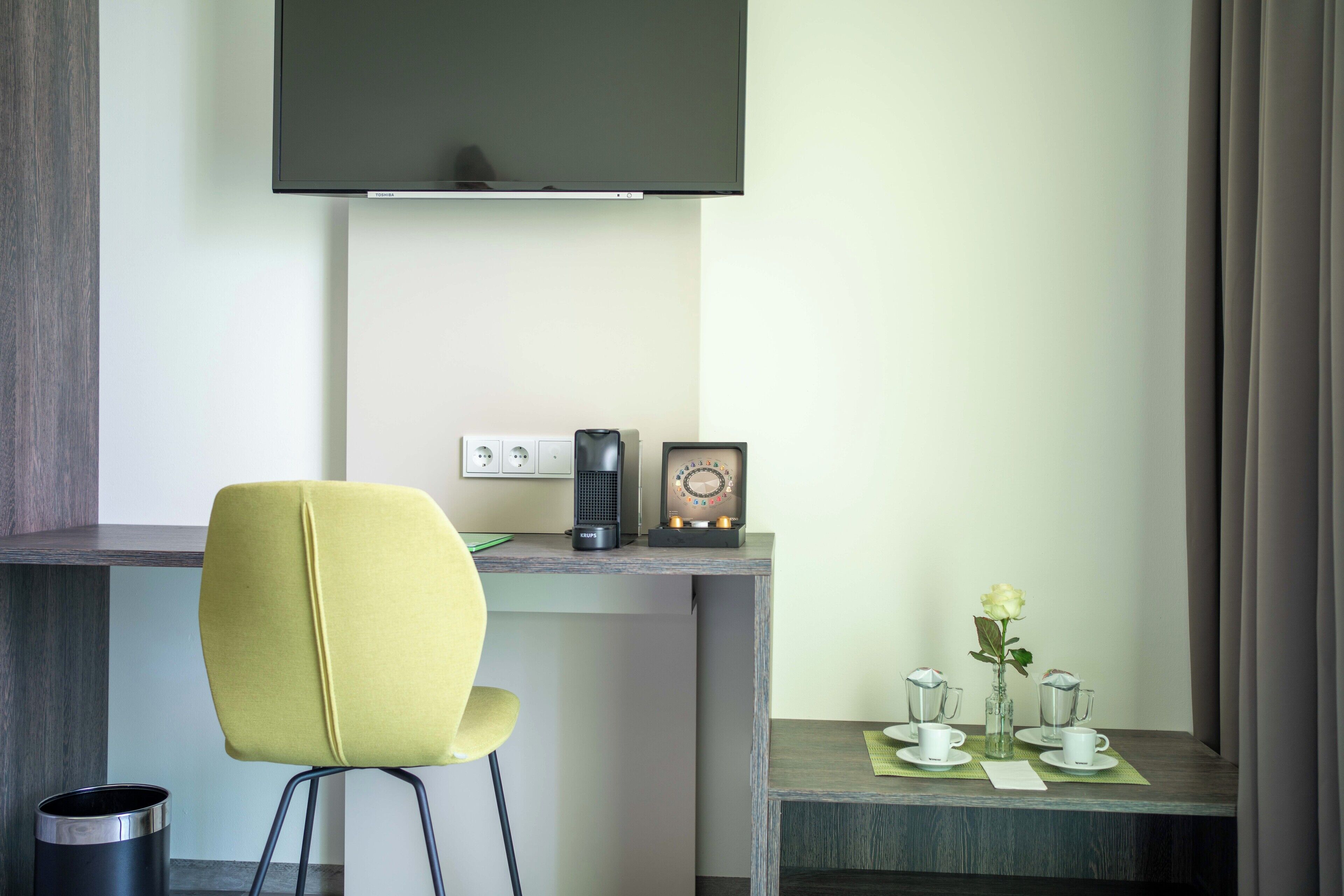 A modern hotel room with a desk, a chair, a coffee maker, a vintage radio, and a small table with a vase of flowers.