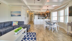 A modern living room with a gray sofa, a white coffee table, and a blue patterned rug. There is a kitchen area with white cabinets and a dining table with white chairs.
