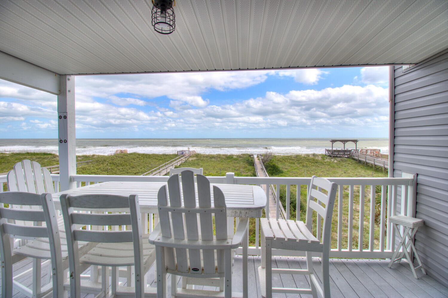 A covered outdoor seating area with white chairs and a table, overlooking a beach with a boardwalk and a pier.